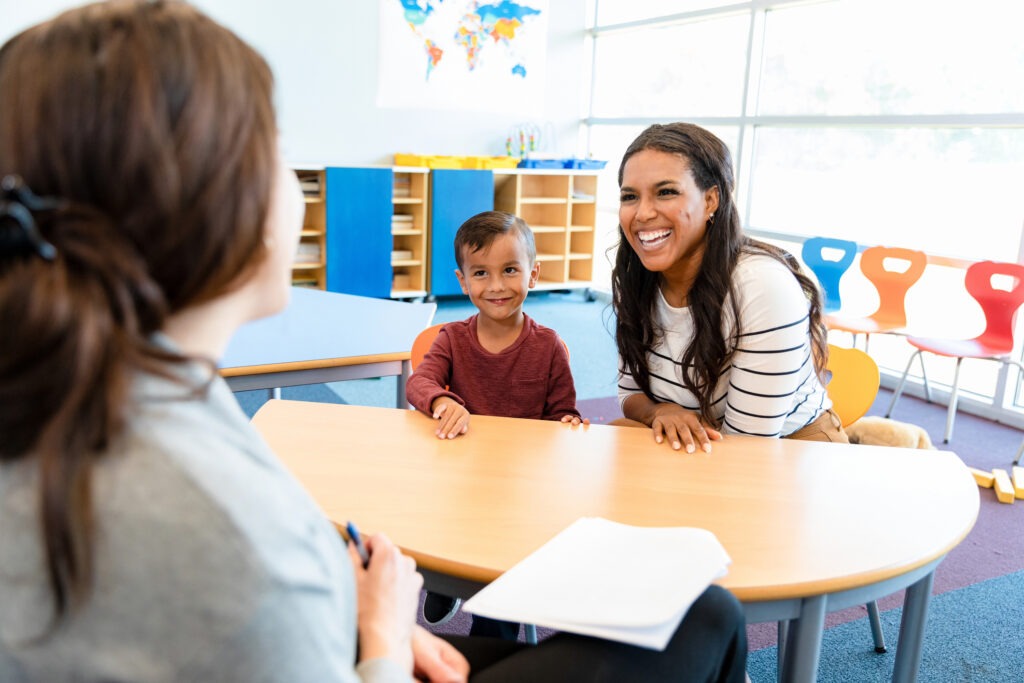 The cute preschool age boy watches quietly as his mid adult mom and his unrecognizable female teacher smile and laugh during the parent teacher conference.