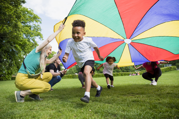 Children playing with a parachute at school during outdoor time. A boy and girl are running underneath it.