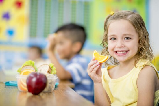 A young girl is sitting indoors in her elementary school classroom. She is smiling at the camera while eating an orange.