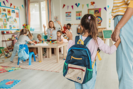 Preschool teacher in kindergarten classroom greeting new student, helping to easily assimilate with others.