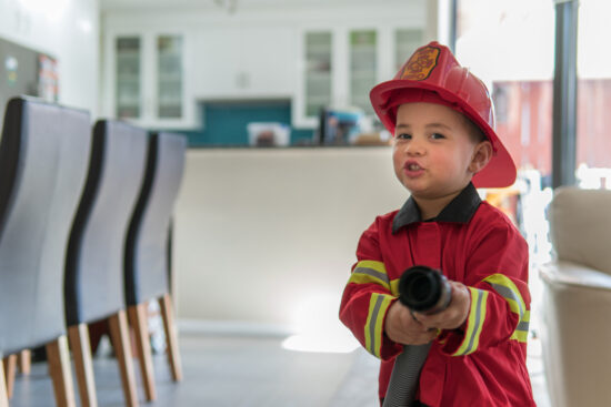 Preschool boy in firefighter costume holding water hose, practicing child safety.