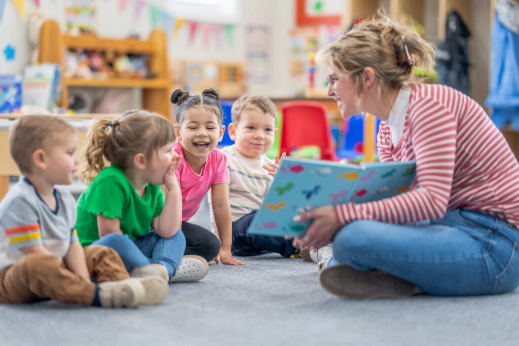 A preschool teacher sits on the floor of her classroom with a small group of students as she reads them a book. The children are each dressed casually and are focused on the story.