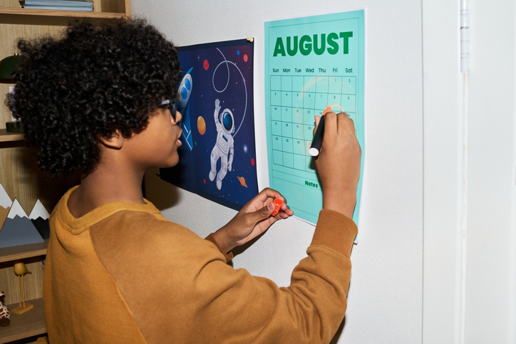 Black child marking August calendar on wall, planning schedule and tracking dates at home. Concept of time management and routine, useful for education or family organization