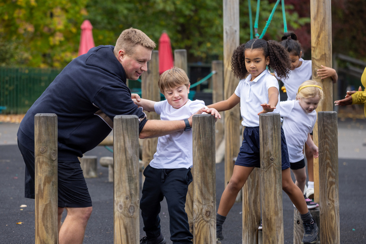 A wide shot two PE teachers supervise young students navigating an outdoor obstacle course. A child with down syndrome balances on wooden posts while one instructor offers support and the others watch from behind. The activity promotes confidence, coordination, and physical skills.