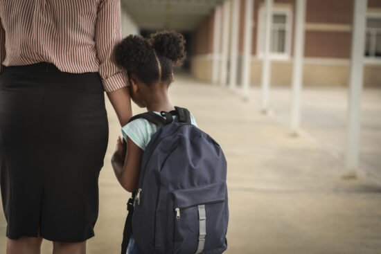 Elementary age, African American girl holds mom or teacher's hand before school begins. She wears a backpack and clings to mom with uncertainty about starting school.