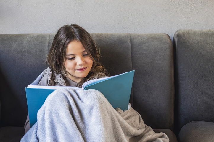 Young girl immersed in reading a book while sitting on a cozy sofa, wrapped in a soft blanket, creating a warm and relaxing atmosphere perfect for quiet moments at home