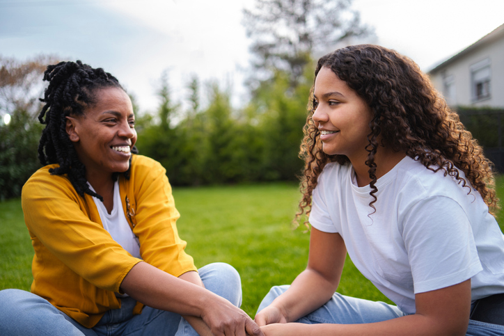 Mother and daughter sitting in grass, holding hands, smiling and talking