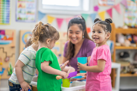 A preschool teacher kneels down with a small group of children as they gather around an activity table filled with rice. They are each dressed casually and are smiling as they enjoy playing together.