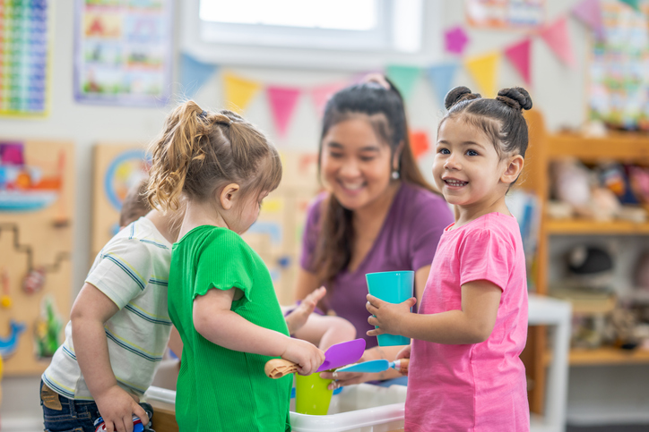 A preschool teacher kneels down with a small group of children as they gather around an activity table filled with rice. They are each dressed casually and are smiling as they enjoy playing together.