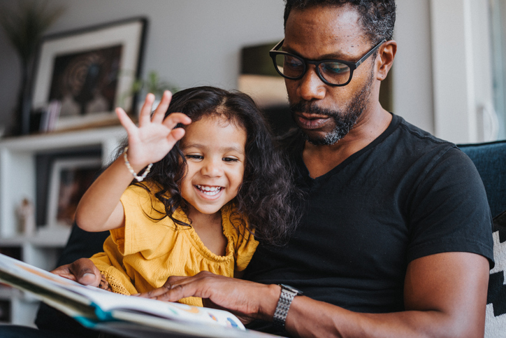 A loving father of African descent sits on the couch in the living room of his home and reads a picture book to his preschool age daughter, who is sitting on his lap smiling.