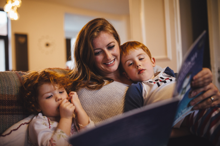 A young family are all sitting together on the sofa in their home, mum is reading a book to her two small children before bedtime.